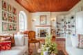 Cozy living room with wooden ceiling, colorful artwork, a fireplace, bookshelves, and vintage furniture.