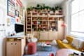 Cozy living room with a yellow sofa, wooden shelving filled with books and plants, and a pink ottoman.