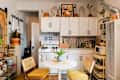 Cozy kitchen with white cabinets, blue patterned tiles, a round table, yellow chairs, and potted plants.