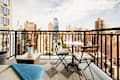 Balcony with a small round table, two chairs, a wooden coffee table, and flowers, overlooking a city skyline.