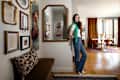 A woman in a green sweater and jeans stands beside a mirror in a stylish living space with framed art and a dining area.