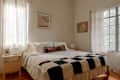 Cozy bedroom featuring a bed with a patterned throw, wooden nightstand, and large windows with sheer curtains.