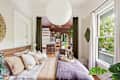 Cozy bedroom with a wooden bed, decorative pillows, and a round paper lantern, leading to a bright living area with plants.