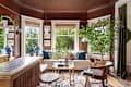 Cozy living room with brown walls, large windows, plants, a beige sofa, colorful cushions, and decorative vases.