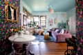 Brightly decorated living room with floral wallpaper, a purple sofa, a round dining table, and colorful artwork.