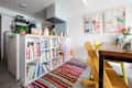 Colorful children's books on a white shelf, striped rug, wooden table with yellow chairs, and abstract wall art.