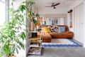 Cozy living room with a brown sectional sofa, colorful cushions, a large plant, and a decorative shelf with books and decor.