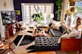 Modern living room with a black leather sectional, wooden coffee table, decorative pillows, and large plants by the window.
