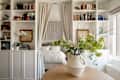 Cozy room with a bed behind curtains, surrounded by bookshelves, a round wooden table, and a floral arrangement.