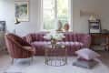 Pink velvet sofa and chair in a living room with a round glass table, floral arrangement, and framed artwork on the wall.