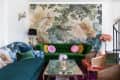 Eclectic living room with green velvet sofa, floral wallpaper, glass coffee table, and colorful cushions.