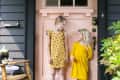 Two children in yellow dresses stand barefoot on a doormat in front of a pink door, with a wooden chair and potted plant nearby.