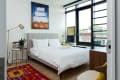 Modern bedroom with a gray upholstered bed, colorful rug, abstract art, and large windows overlooking rooftops.