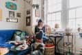 Cozy living room with a woman knitting on a wicker chair, surrounded by colorful decor, plants, and a cat.