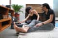 Couple sitting on a rug, listening to a record player, with a wooden shelf and potted plant in the background.