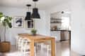 Dining room with wooden table, white chairs, black pendant lights, and a view into a kitchen with open shelving.