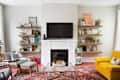 Living room with a wall-mounted TV, floating shelves, yellow sofa, gray chairs, and a patterned rug.