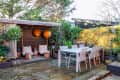 Outdoor patio with wooden deck, dining table, chairs, yellow wall, plants, and two orange lanterns hanging in a cozy seating area.