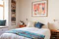 Cozy bedroom with a white bed, colorful throw, abstract art, and built-in bookshelf by a window seat.