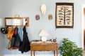 Wooden console table with lamp, plant, and decor under a mirror and colorful woven wall hanging. Coats and bags hang nearby.