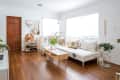 Living room with white sofa, wooden coffee table, plants, shoe rack, and decorative pillows on wooden flooring.