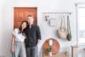 Couple smiling in front of a wooden door, with wall hooks holding bags and decor items in a bright room.