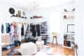 Bright walk-in closet with hanging clothes, shoe racks, hats, and bags on shelves, featuring a white pouf and wooden floor.