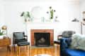 Living room with brick fireplace, blue sofa, wooden chairs, and plants on mantel. Mid-century modern decor and hardwood floor.