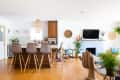 Open-plan kitchen and living room with bar stools, wicker chairs, plants, and a TV above a patterned fireplace.