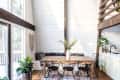 Dining area with wooden table, black chairs, white vase with greenery, macramé wall hanging, and large windows.