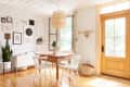 Dining area with wooden table, white chairs, wall art, potted plant, and wicker pendant light near a wooden door.