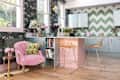 Colorful kitchen with green chevron tiles, pink island, gold wire chairs, and floral wallpaper.