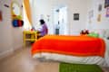 Child sitting at a yellow desk in a bedroom with an orange bedspread, colorful wall art, and a green rug.