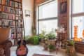 Cozy living room with a leather chair, guitar, tall bookshelf, potted plants, and a bar cart by a large window.