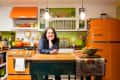 Woman leaning on a wooden kitchen island with orange appliances and green walls.