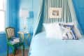 A bed with a blue bedspread that matches the walls featuring a pillow with painting on it.