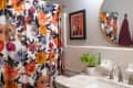 Bathroom with floral shower curtain, round mirror, framed art, and potted plant on marble countertop.