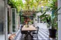 Dining area with wooden table, black chairs, lush plants, and a dog on checkered floor under a vine-covered ceiling.