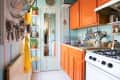 Narrow kitchen with orange cabinets, striped walls, hanging utensils, and a mirror door reflecting a hallway.