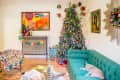 Festive living room with a decorated Christmas tree, colorful art, a "Merry + Bright" banner, and a cat on a teal sofa.