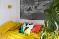 Yellow bed with colorful geometric pillows, large abstract wall art, and a green monstera plant.