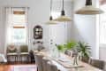 Dining room with a long wooden table, beige chairs, pendant lights, and green plants in glass vases.
