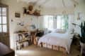 Cozy bedroom with white bedding, wooden shelves, books, plants, and a large window with sheer curtains.