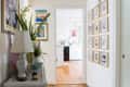 Hallway with framed art on walls, plants on a console table, leading to a bright kitchen with wooden floors.