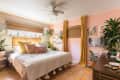 Cozy bedroom with pink walls, plants, a bed with colorful pillows, and yellow curtains. Art decorates the walls.
