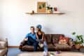 Couple sitting on a brown leather sofa with colorful pillows, plants, and a shelf with decor above.