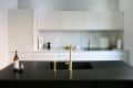 Modern kitchen with white cabinets, marble backsplash, black countertop, gold faucet, wine bottle, and glass.