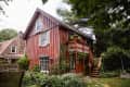 Red wooden house with white shutters, surrounded by lush greenery and a small garden, featuring a wooden staircase.