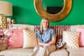 Woman sitting on a beige sofa with pink and gold polka dot pillows, green wall, and round gold mirror.