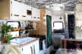 Camper interior with patterned ceiling, white cabinets, plants, geometric backsplash, and cozy bed with patterned bedding.